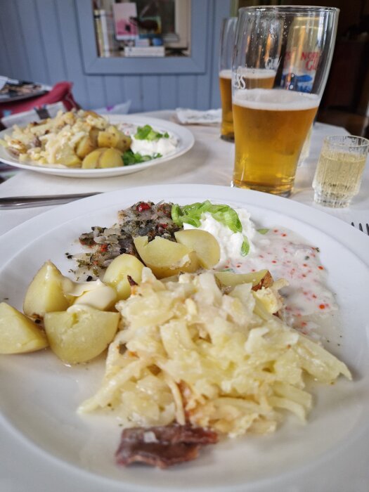 Tallrik med löksill med chili, potatis, gräddfil, salladslök och Jansson. Bakom står glas med öl och snaps.