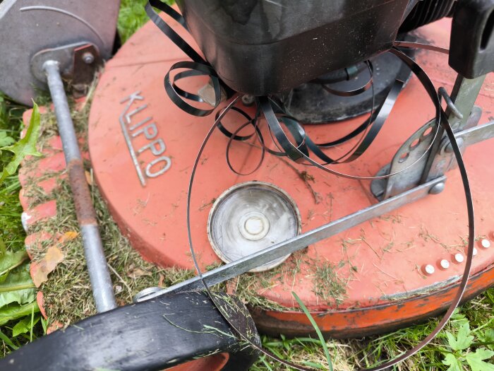 Trasig gräsklippare med avbrutna remmar vid klippning, gräs fastnat under röd kåpa med "Klippo" tryckt på.