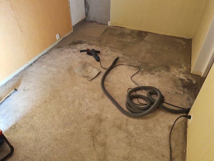 Rum med synligt betonggolv, en elektrisk slipmaskin och dammsugarens slang spridd på golvet, under pågående renovering.