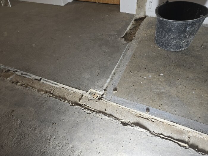 Betonggolv med en upptagen ränna som avslöjar trä inuti. Byggarbetsyta med hink synlig i högra hörnet.