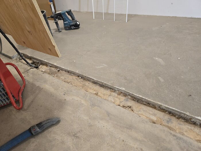 En ränna i ett betonggolv visar borttaget material, med verktyg och träskiva i bakgrunden, troligen från renovering av källare.