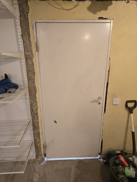 Vit dörr i källare, omgiven av gul vägg och hylla till vänster. Verktyg och hink på golvet. Dörren har ojämn passform, med en synlig springa nederst.