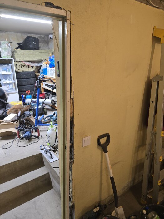 Öppen dörrkarm i en källarvägg under renovering. Verktyg och byggmaterial syns i rummet bakom. En spade och en förstaplanslaser finns i bild.