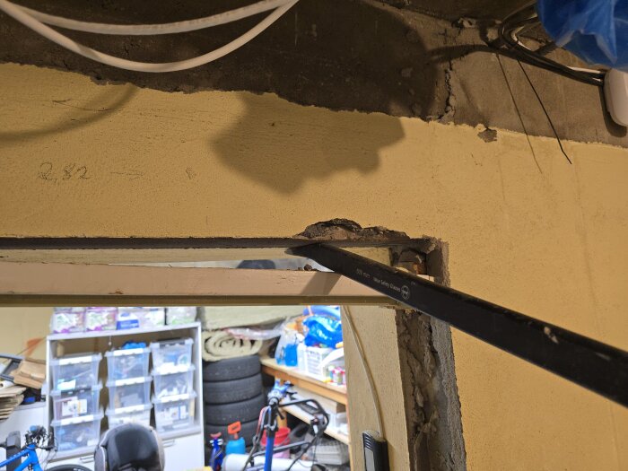 Verktyg används för att öppna upp en dörröppning. Borttagen dörrkarm med synligt armeringsnät i korridor under renovering. Kartonger och cykel syns i rummet.