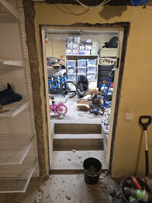 Vy från en källare med en öppen dörr, trappa ner till ett garage med cyklar och förvaringslådor, omgiven av byggmaterial och verktyg.