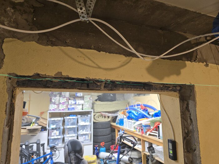 Öppning i vägg mellan garage och källare under renovering, med kablar och verktyg synliga. Inredning och diverse föremål i bakgrunden.