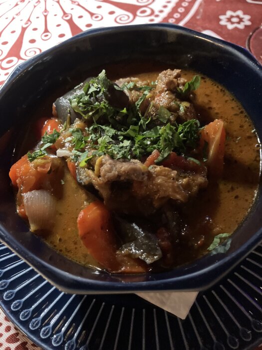 En skÄl med georgisk gryta innehÄllande kött, tomater, lök, aubergine och örter, serverad pÄ en dekorativ röd och vit duk.