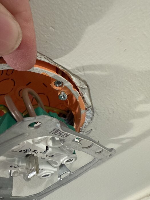 Närbild på en elektrisk kopplingsdosa i en vägg med skruvar och exponerade kablar, där en hand pekar mot en monterad komponent.