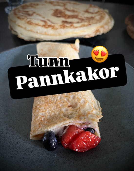 Tunn pannkaka fylld med fruktsallad och grädde på en tallrik, med staplade pannkakor i bakgrunden.