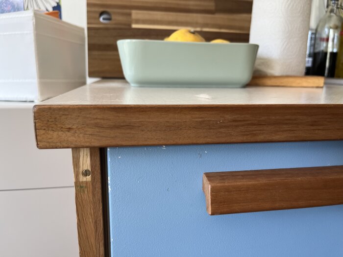 Köksbänk med brun kantlist och blålackerad lådfront, en fruktskål och hushållspappershållare på bänkskivan i bakgrunden.