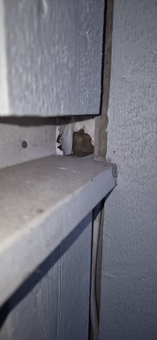 Hål i skyddsplast mellan två ytterväggar i ett hörn, där träpanel saknas. Plasten är trasig och hörnet skyddas delvis av ett utskjutande tak.