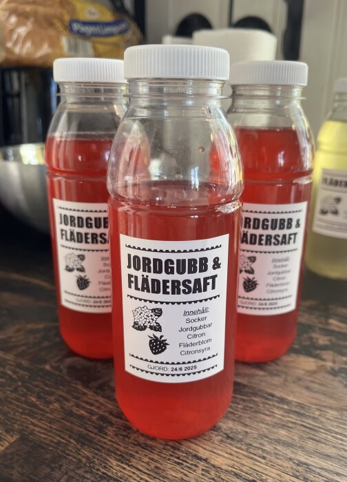 Flaskor med jordgubb och flädersaft på ett bord, etiketterade med innehållsförteckning och produktionsdatum.