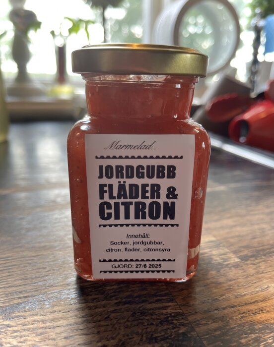 Burk med jordgubb, fläder och citronmarmelad på ett bord, etikett med innehållsförteckning och tillverkningsdatum 27/6 2025.