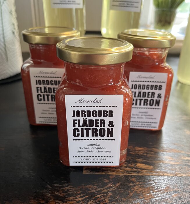 Burkar med jordgubb, fläder och citronmarmelad står på bordet, omgivna av flaskor med flädersaft.