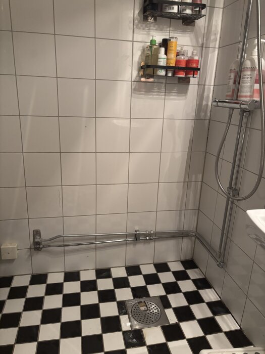 Dusch med vitkaklade väggar, hyllor med flaskor, och schackrutigt svartvitt kakelgolv med golvbrunn.
