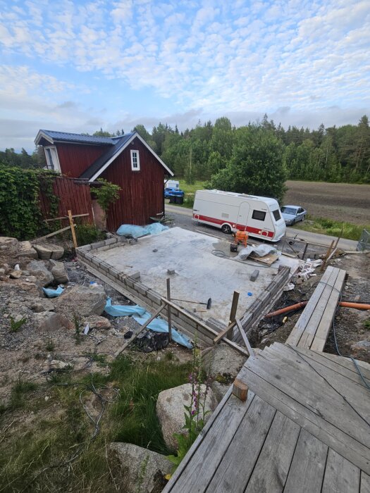 Byggarbetsplats för carport med förråd i sutteräng, med brädor, betongfundament och verktyg. Röd byggnad och skog i bakgrunden.