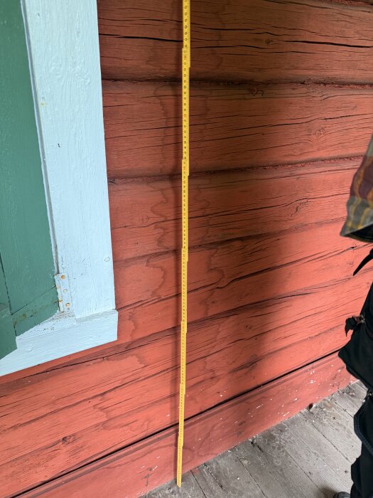 Röd träpanel på torpvägg med måttband lutat mot väggen vid ett fönster med grön fönsterkarm.