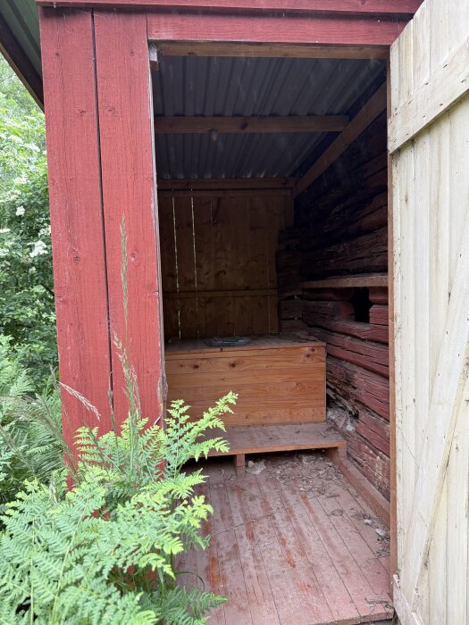 Halvöppet gammalt utedass med rött träpanel, träbänk och plåttak; omgiven av grönska och behöver renovering.