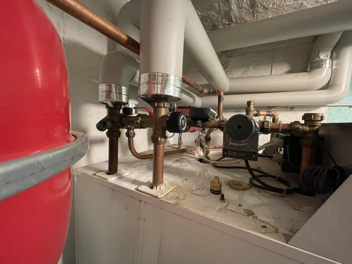 Rör och ventiler ovanpå en central med kopparledningar och ett rött tryckkärl synliga i ett maskinrum.