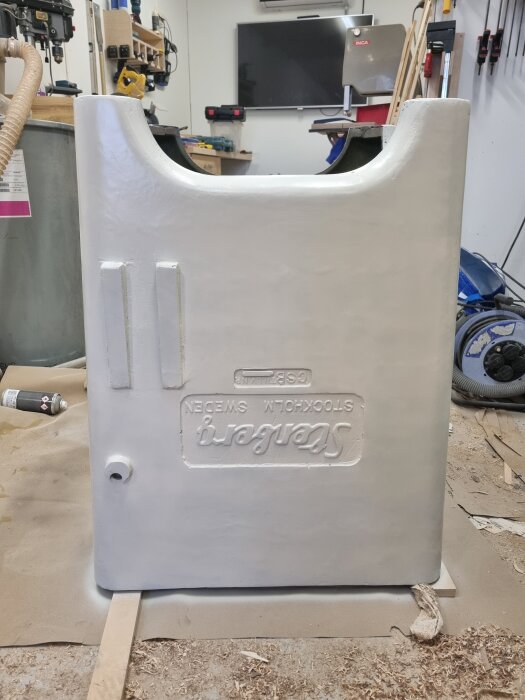 Krämvit målad sågkropp med slät yta, omgiven av verktyg och sprayfärgsburkar i en snickarverkstad.