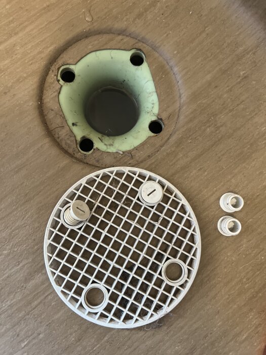 Rördelsbrunn med avtagbart galler och fyra skruvhål, samt två lösa pluggformade delar på trägolv.