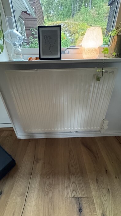 Väggmonterat 400V-element under ett fönster med dekorativa föremål på fönsterbrädan, inkluderar en lampa och en krukväxt.