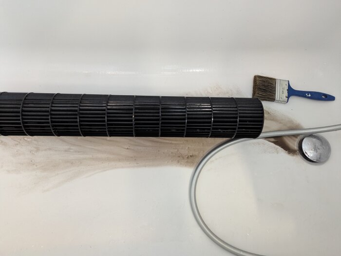 Rengjord trumma från en luftvärmepump med smutsrester på vit yta, en pensel och en slang syns i bild.