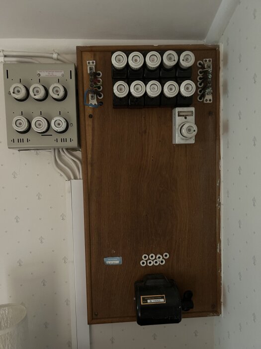 Elcentral med porslinssäkringar och träpanel i äldre byggnad. Svarta kablar och ledningar synliga ovanför säkringspanelen.