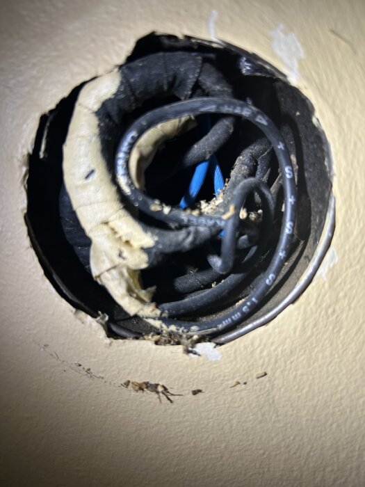 Gamla tygisolerade ledningar och kablar i ett cirkulärt hål i vägg, med trasig tapet och smuts runt kanterna.