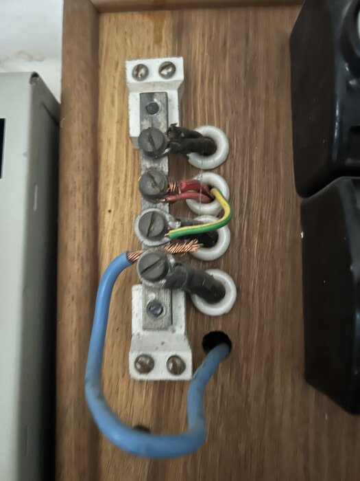 Gammalt elsystem med säkringar och ledningar i ett träskåp, inklusive en blå och flera färgade kablar.