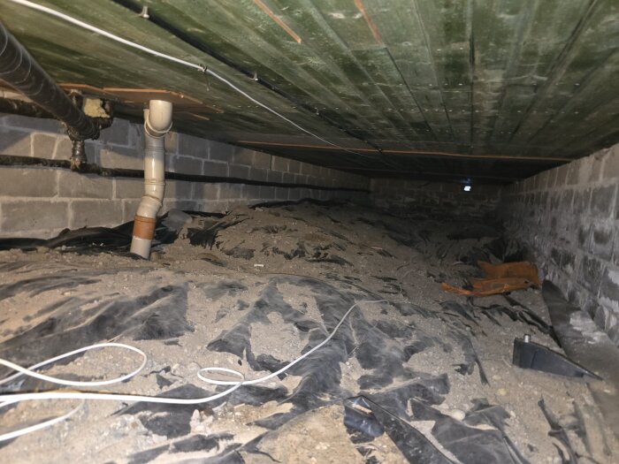 Krypgrund med sand och rörledningar, betongtak syns ovanför.