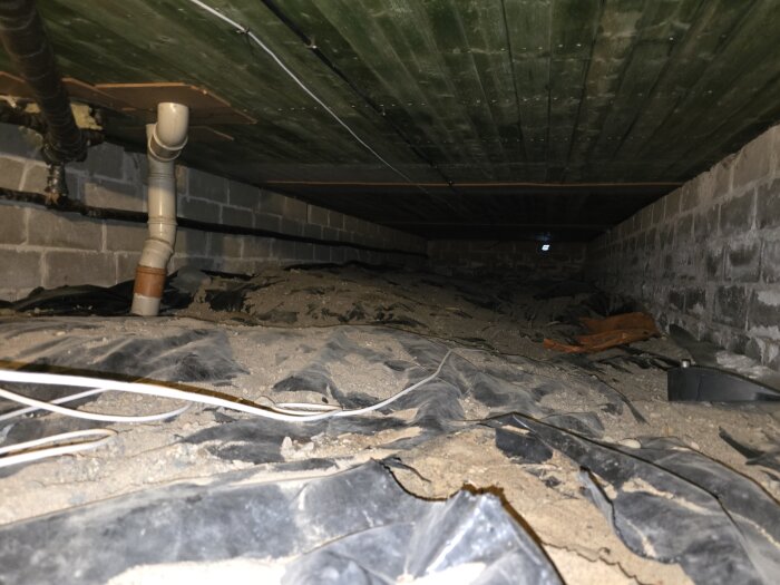 Bild av krypgrund med sand och isoleringsplast under ett hus, med del av rörsystem synligt.