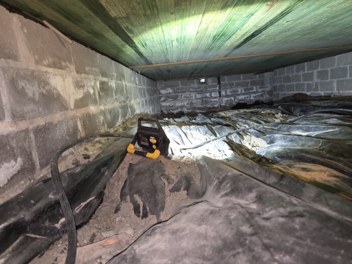 Under huset visar bild en grov sandig yta med plastfolie, murad vägg och arbetslampa. Fråga om att ta bort sand inför isoleringsarbete.