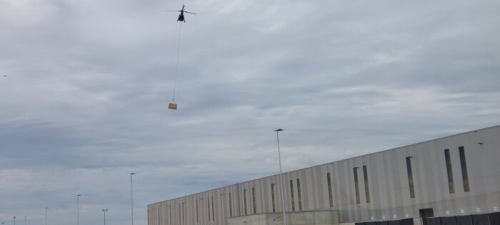 Helikopter lyfter en last ovanför ett stort industribyggnadstak under molnig himmel.
