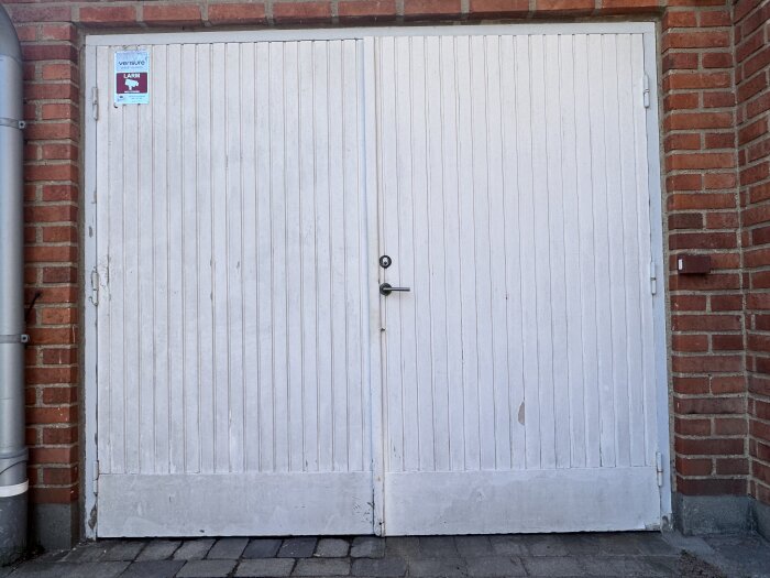 Vit trädörr till garage, inramad av tegelvägg. Handtag i mitten, något sliten yta med färg som flagnar, sidoskylt om larm.