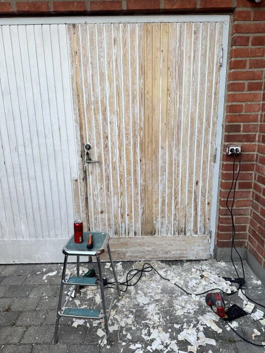 Garageport under restaurering, trärent synligt där linoljefärg är bortskrapad. Verktyg och burk på pall, färgrester på marken.