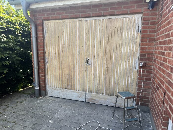 Gammal garageport med bortskrapad linoljefärg avslöjar trärent trä, med stegpall och kabel på stenläggning framför tegelvägg.
