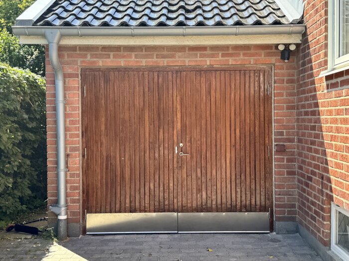 Renoverad garageport i trä med vertikala träpaneler, omgiven av tegelväggar och ett tak med svarta takpannor.