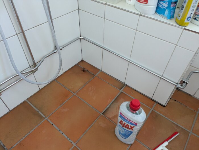 Fläckig klinkergolv vid badkarrens tidigare plats med städpå produkter, inklusive Ajax-behållare, redo för rengöring.
