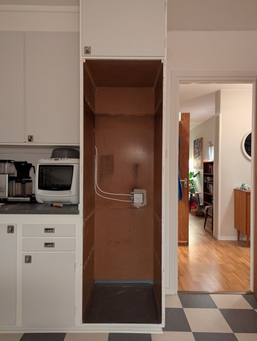 Tomt högskåp i kök, delvis demonterat, med synliga elkablar och vägg bakom. Köksinredning med vitvaror till vänster.