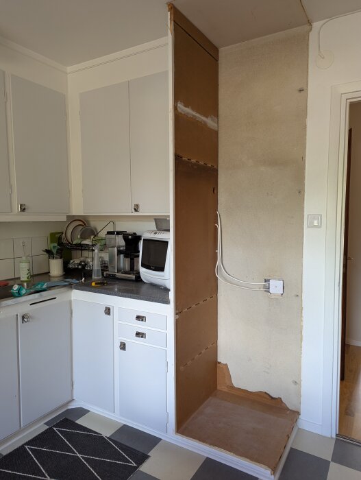 Tom nisch i kök med borttagen del av högskåp, synliga kablar och vägg bakom; mikrovågsugn på bänkskiva bredvid.