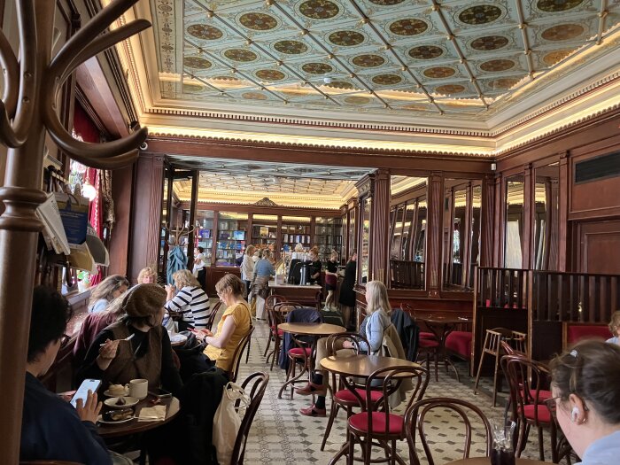 Ett elegant kafé med dekorativt tak, speglar och röda stolar, där människor sitter vid små bord, vissa med telefoner och kaffe.