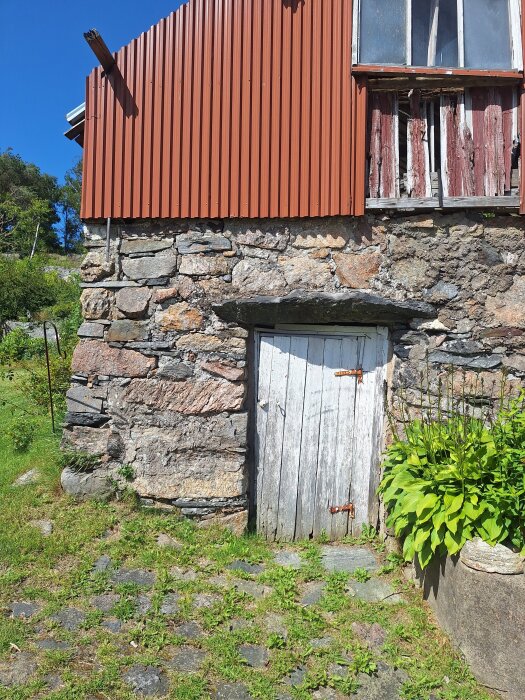 Stenbyggd jordkällare i sluttning med en trästuga ovanpå. Trästugan har ett rött plåttak och nedre delen visar en sliten dörr.