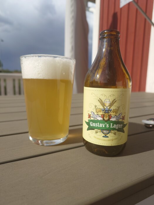 Glas med skummande öl bredvid en flaska "Gustav's Lager" på ett bord, med ett rött hus i bakgrunden och molnig himmel.