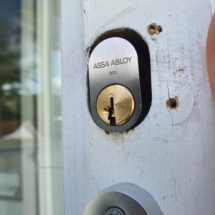 Oval cylinderlås Assa Abloy 1308 i en altandörr, monterad med svarta skruvar, omgiven av hål och skador på dörrkarmen.