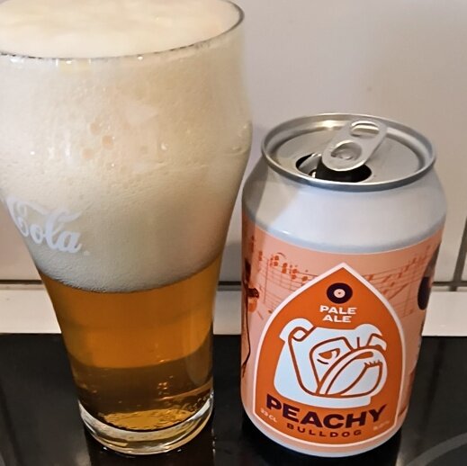 Ett glas med skummande pale ale bredvid en burk "Peachy" från Angry Bulldog, fotograferat på en köksyta.