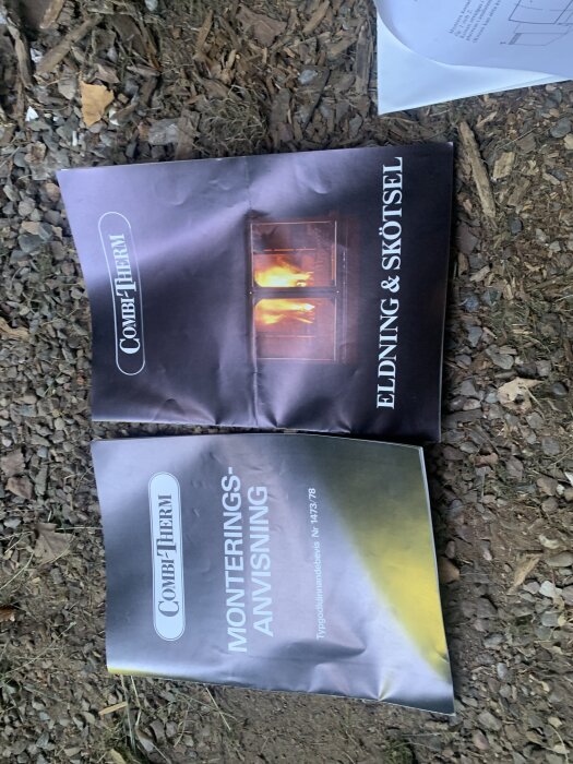 Bruksanvisningar för en Combitherm-kamin ligger på marken, en för eldning och skötsel, den andra för monteringsanvisning.