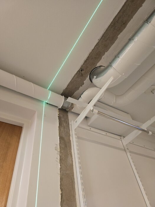 Vägg med borttagen panel visar ränna och ventilationsrör i taket. Grön laserlinje markerar ny väggposition för förråd.