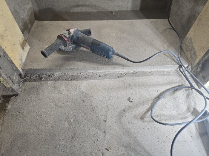 Betonggolv med ränna nära en vägg; en vinkelslip med elsladd ligger på golvet.