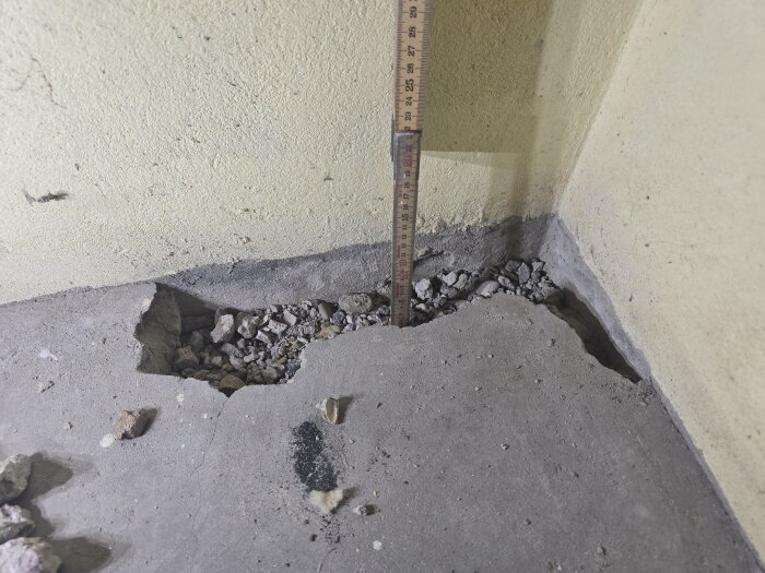 Hörnet i ett rum med knäckt betonggolv och ett mätverktyg som visar djupet av en ränna fylld med stenar och grus mot en gul vägg.